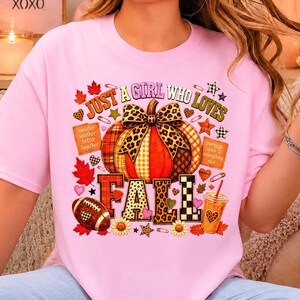 May include: A light pink t-shirt featuring a graphic print of a pumpkin with a leopard print bow, surrounded by autumn leaves, a football, a drink, and the text "JUST A GIRL WHO LOVES FALL" along with "sweater weather better together" and "pumpkin spice & everything nice."