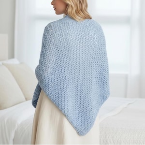 May include: A light blue, open-weave shawl. The shawl is draped over the shoulders and arms. The person is wearing a cream-colored dress. The background includes a bed with white linens and pillows.
