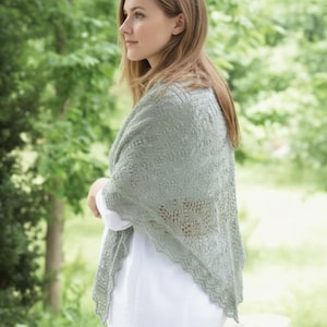 May include: A pale green shawl with a delicate lace pattern is draped over the shoulders. The shawl has a scalloped edge and is worn over a white top. The background is a soft focus of green foliage.