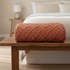 Peut inclure: Une couverture tissée de couleur terre cuite est posée sur un banc en bois. La couverture présente un motif de tissage épais et texturé. La chambre comprend un lit avec une literie aux tons neutres et une table de chevet avec une lampe.