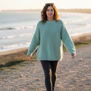 Op de afbeelding: Een persoon draagt een lichtblauwe gebreide trui en een donkergrijze legging en loopt op een pad in de buurt van de zee. Een zonnebril ligt op de trui. De trui heeft een geribbelde textuur en lange mouwen.