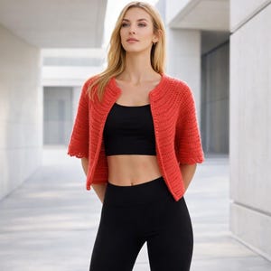 May include: A coral-colored, short-sleeved, crocheted bolero jacket. The open-front cardigan has a scalloped edge and is worn over a black crop top and black leggings. The jacket is a vibrant, handmade piece.