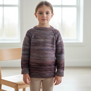 May include: A child wearing a knitted sweater with a raglan sleeve design. The sweater features a color gradient, transitioning from dark burgundy at the bottom to lighter shades of purple and grey at the top. The child is wearing beige pants.