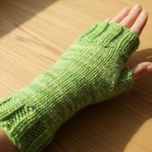 Könnte beinhalten: Ein grüner Strick-Fingerhandschuh mit geripptem Bündchen und strukturiertem Maschenmuster. Der Handschuh ist aus weichem, hellgrünem Garn mit weißen Flecken gefertigt. Der Handschuh wird an einer Hand gezeigt.
