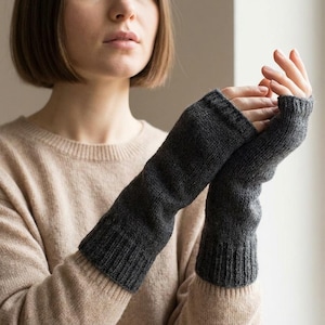 Op de afbeelding: Donkergrijze gebreide vingerloze handschoenen. De handschoenen zijn lang, reiken tot aan de onderarm en hebben een geribbelde manchet. Het materiaal lijkt zacht en warm, geschikt voor koud weer. De handschoenen worden tegen een neutrale achtergrond getoond.