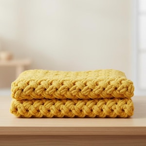 May include: Two folded mustard yellow blankets with a thick, woven texture are stacked on a light wood surface. The blankets have a repeating pattern of large, rounded knots. The background is blurred.