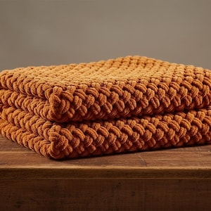 May include: A folded, chunky knit blanket in a warm, earthy orange hue. The blanket is made of thick, soft yarn, and the texture is visible. The blanket is folded neatly on a wooden surface.