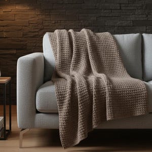 May include: A taupe-colored waffle-knit throw blanket draped over a light gray sofa. The blanket's texture is visible, and it contrasts with the smooth fabric of the couch. The scene is set in a well-lit room with a modern aesthetic.