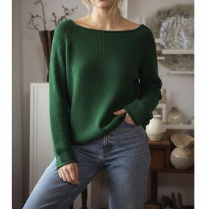 May include: A forest green sweater with a wide boat neck and long sleeves. The sweater is paired with light blue denim jeans. The model is standing in front of a neutral background with decorative items.