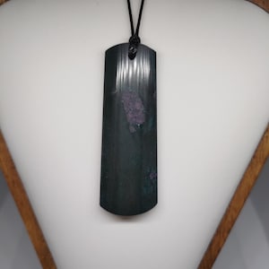 May include: A dark green rectangular pendant with a black cord. The pendant has a curved base and a textured surface with purple and green accents. The pendant is displayed on a white surface.