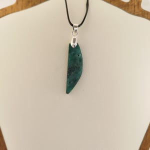 May include: A pendant necklace with a teal-coloured, semi-circular stone. The stone is suspended from a silver-coloured metal clasp and a black cord. The stone has dark green and black inclusions. The necklace is displayed on a white surface.