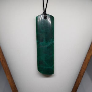 May include: A dark green rectangular pendant with a rounded base hangs from a black cord. The pendant has a marbled effect, with lighter green swirls. The necklace is displayed on a white surface.