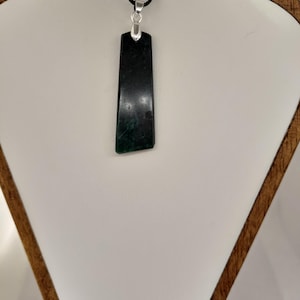 May include: A dark green rectangular pendant with a silver-coloured bail hangs from a black cord. The pendant has a polished, smooth surface and a slightly irregular bottom edge. The necklace is displayed on a white surface.