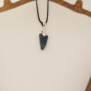 May include: A dark teal pendant necklace with a silver-coloured bail and a black cord. The pendant is a triangular shape with a rounded top. The necklace is displayed on a white surface.