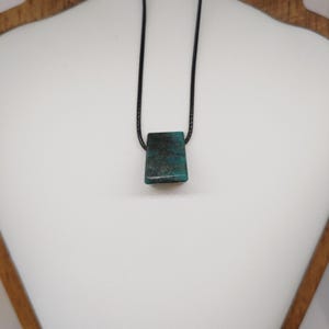 May include: A rectangular pendant necklace with a teal and black stone. The stone is suspended from a black cord. The necklace is displayed on a white mannequin bust. The pendant is approximately 2.5 cm in length.