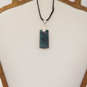 May include: A rectangular pendant with a dark teal, textured surface hangs from a black cord. The pendant is attached to a silver-coloured bail. The necklace is displayed on a white surface.