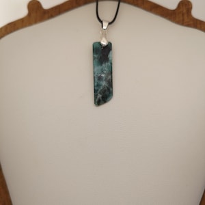 May include: A rectangular pendant with a dark teal and black marbled pattern hangs from a black cord. The pendant is attached to a silver-coloured bail. The necklace is displayed on a white surface.