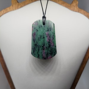 Peut inclure: Un collier pendentif rectangulaire avec une pierre naturelle. La pierre présente une couleur de base verte avec des inclusions rouges et noires. Le pendentif est suspendu à un cordon noir. Le collier est présenté sur une surface blanche.