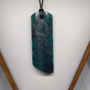 May include: A rectangular pendant with a dark teal and blue marbled pattern, suspended from a black cord. The pendant has a rounded bottom edge and a hole at the top for the cord. The background is a neutral white.