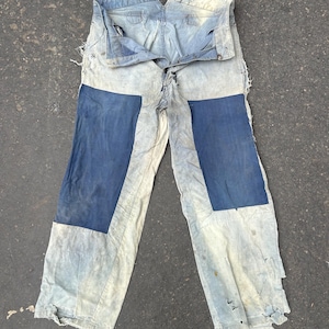 Peut inclure: Pantalon de travail en jean bleu clair avec deux grands patchs bleus sur le devant. Le pantalon présente des signes d'usure, avec des bords effilochés et des zones abîmées. La taille est fermée par un bouton et une fermeture éclair.