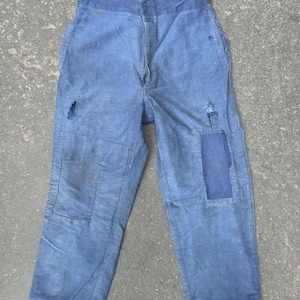 May include: Faded blue work pants with patches and visible wear. The pants have a high waist and a button fly. Patches are sewn onto the knees and thigh. The fabric appears to be a durable cotton or linen blend.