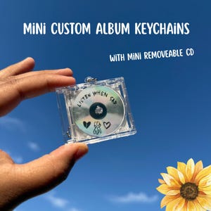 May include: A clear plastic square keychain case containing a removable mini CD. The CD features the text LISTEN WHEN SAD, a broken heart icon, a small figure, and a heart icon. The text MINI CUSTOM ALBUM KEYCHAINS WITH MINI REMOVEABLE CD is visible.