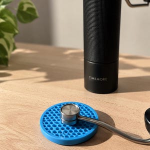 Peut inclure: Un moulin à café manuel cylindrique noir avec une prise texturée et le nom de marque TIMEMORE imprimé sur la base. Un tapis en silicone circulaire bleu avec un motif en nid d'abeille est posé devant, supportant une manivelle en métal argenté.