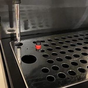 Peut inclure: Gros plan sur une machine à expresso noire et argentée. La machine est dotée d'un bac d'égouttement en acier inoxydable avec de multiples trous. Un bouchon rouge et noir est visible sur le bac. Un bec verseur argenté est positionné au-dessus du bac.