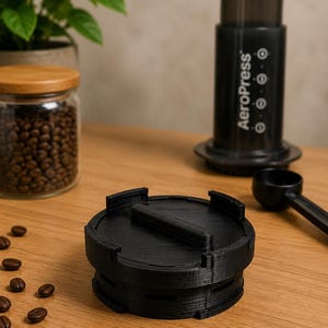 以下が含まれることがあります： 中央にハンドルがあり、縁に切り込みが入った黒い円形の3Dプリント製コーヒーフィルターホルダー。近くには黒いエアロプレスコーヒーメーカー、黒いプラスチック製のスプーン、焙煎したコーヒー豆が入ったガラス瓶が置かれています。