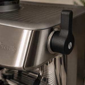 以下が含まれることがあります： つや消しステンレススチール製のBrevilleエスプレッソマシン。側面には白いコーヒー豆のアイコンが付いた黒いスチームワンドレバーが取り付けられています。マシンにはデジタルディスプレイ画面、上部のウォーミングトレイ、金属製のフロントパネルが備わっています。