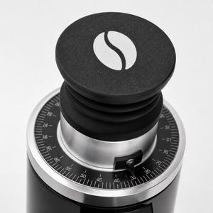 Peut inclure: Un moulin à café noir avec un cadran argenté et un dessus noir orné d'un logo de grain de café blanc. Le cadran comporte des chiffres et des marquages pour un broyage précis. Le design du moulin est élégant et moderne.