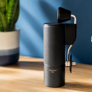 May include: A black cylindrical manual coffee grinder with a textured grip and a metal handle featuring a black knob. The device is labeled with the text CHESTNUT C3s on the lower body. It has a sleek, vertical design with a metallic crank arm.