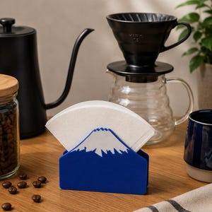 Op de afbeelding: Een blauwe koffiefilterhouder in de vorm van de berg Fuji, met daarin een stapel witte papieren koffiefilters. De houder heeft een gestileerd ontwerp van een bergtop met een wit bovendeel en een stevige, stabiele blauwe basis.