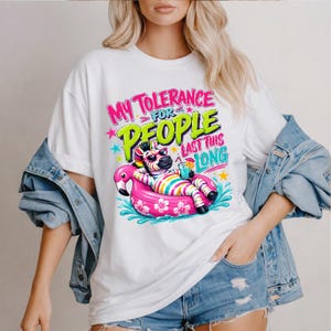 Può includere: T-shirt bianco con una grafica colorata di una zebra con occhiali da sole, che si rilassa su un galleggiante a forma di fenicottero rosa. Il testo recita "My Tolerance For People Last This Long" in colori vivaci. La maglietta ha una vestibilità ampia.
