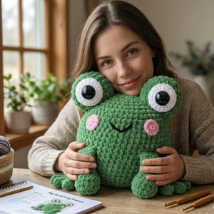 May include: A large, green crocheted frog with big, white-rimmed eyes and pink cheeks. The frog is being held. A pattern for the frog is on the table. Crochet hooks are also on the table.
