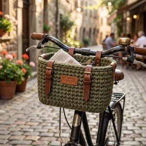 May include: A dark olive green crocheted bicycle basket attached to black handlebars. The basket features two brown leather straps with metal buckles and a rectangular leather label reading OLIVE ADVENTURE Crochet Bike Basket EST 1993.