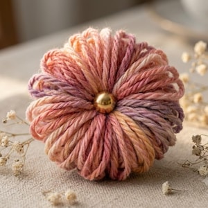 May include: A round, decorative object crafted from variegated yarn in shades of pink, orange, and purple, featuring a smooth, polished gold-colored metal bead centered in the middle, creating a textured, radial pattern resembling a small flower.