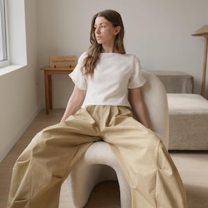 May include: A short-sleeved white top with a boat neckline and flared sleeves paired with wide-leg tan trousers featuring front pleats. The outfit consists of a solid white blouse and loose-fitting beige trousers that reach the floor.