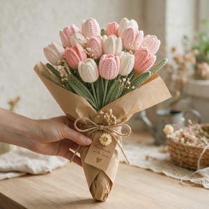 Könnte beinhalten: Ein Strauß aus gehäkelten Tulpen in zartem Rosa und Weiß, ergänzt durch grüne gehäkelte Blätter und kleine Trockenblumen. Die Stiele sind in braunes Papier eingewickelt, das mit einer Schnur und einem Schild mit Handmade with love versehen ist.