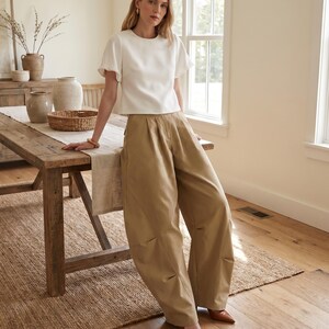 May include: A white short-sleeved blouse with a round neckline and pleated tan wide-leg trousers. The blouse features slightly puffed sleeves, while the trousers have a high-waisted fit with visible pleats at the waist and a loose, flowing silhouette.