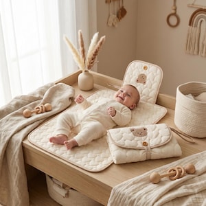 Könnte beinhalten: Ein Baby liegt auf einer cremefarbenen, gesteppten Wickelauflage mit Teddybärenmuster. Eine passende Wickelauflagenabdeckung und eine Windeltasche befinden sich in der Nähe. Eine Holzrassel und ein Weidenkorb sind ebenfalls zu sehen.