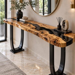 May include: A long console table featuring a live-edge wood slab with a central black resin river inlay. The table is supported by two U-shaped black metal legs. On the surface sits a black vase with greenery, a small bowl, and a reed diffuser.