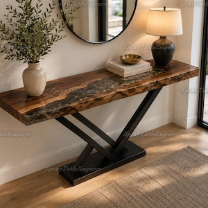 May include: A rectangular console table featuring a natural wood slab with a central dark resin inlay, supported by a black metal V-shaped base. The tabletop displays a live edge finish, while the base rests on a solid rectangular black metal plate.