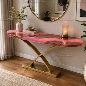 May include: A console table featuring a live-edge wood slab with a translucent pink epoxy resin center, supported by a gold-toned metal base with crossed legs. The table surface holds a white vase, a small bowl, a book, and a glass candle holder.