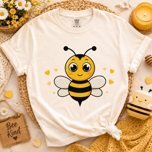 May include: A cream-colored short-sleeve t-shirt featuring a central graphic of a cartoon bee with large eyes, black and yellow stripes, and white wings, surrounded by small yellow hearts. The shirt has a crew neckline and a Comfort Colors label.