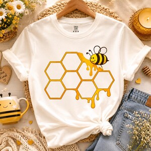 May include: A white short-sleeve t-shirt featuring a graphic print of a yellow and black bee perched on a cluster of orange-outlined hexagonal honeycomb cells with dripping honey. The shirt has a crew neckline and a Comfort Colors brand label.