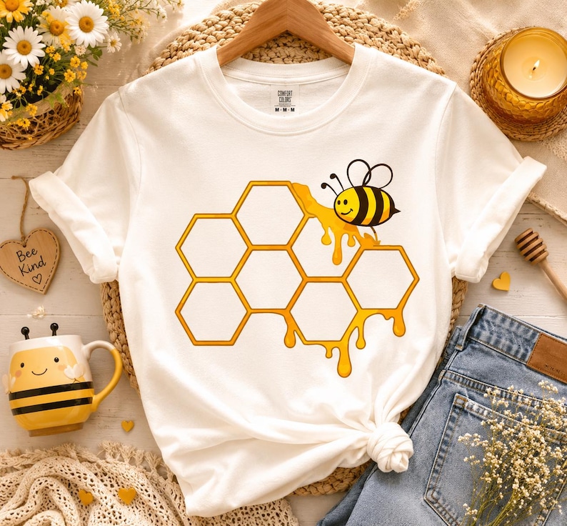 May include: A white short-sleeve t-shirt featuring a graphic print of a yellow and black bee perched on a cluster of orange-outlined hexagonal honeycomb cells with dripping honey. The shirt has a crew neckline and a Comfort Colors brand label.