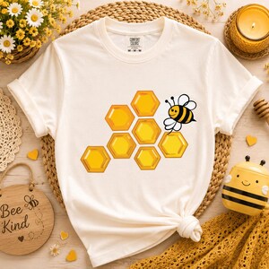May include: A white short-sleeve t-shirt featuring a graphic print of yellow hexagonal honeycomb cells and a small cartoon bee. The shirt has a crew neckline and a Comfort Colors brand label printed on the inside of the collar area.