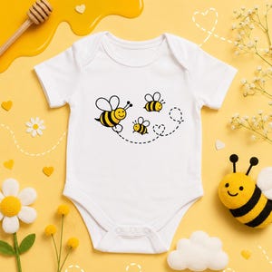 May include: A white short-sleeved baby bodysuit featuring a graphic print of three yellow and black striped bees. The bees are depicted in flight with a dotted line trail that forms a heart shape. The garment has a round neckline and snap closures.