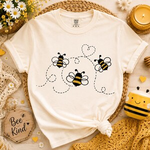 May include: A cream-colored short-sleeve t-shirt featuring a graphic print of three stylized bees with yellow and black striped bodies. The bees are connected by a dashed black line that forms a heart shape above them. The shirt has a crew neckline.